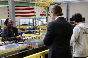 Governor Newsom tours a power factory and shares a smile with a brown skin-toned woman in protective glasses.