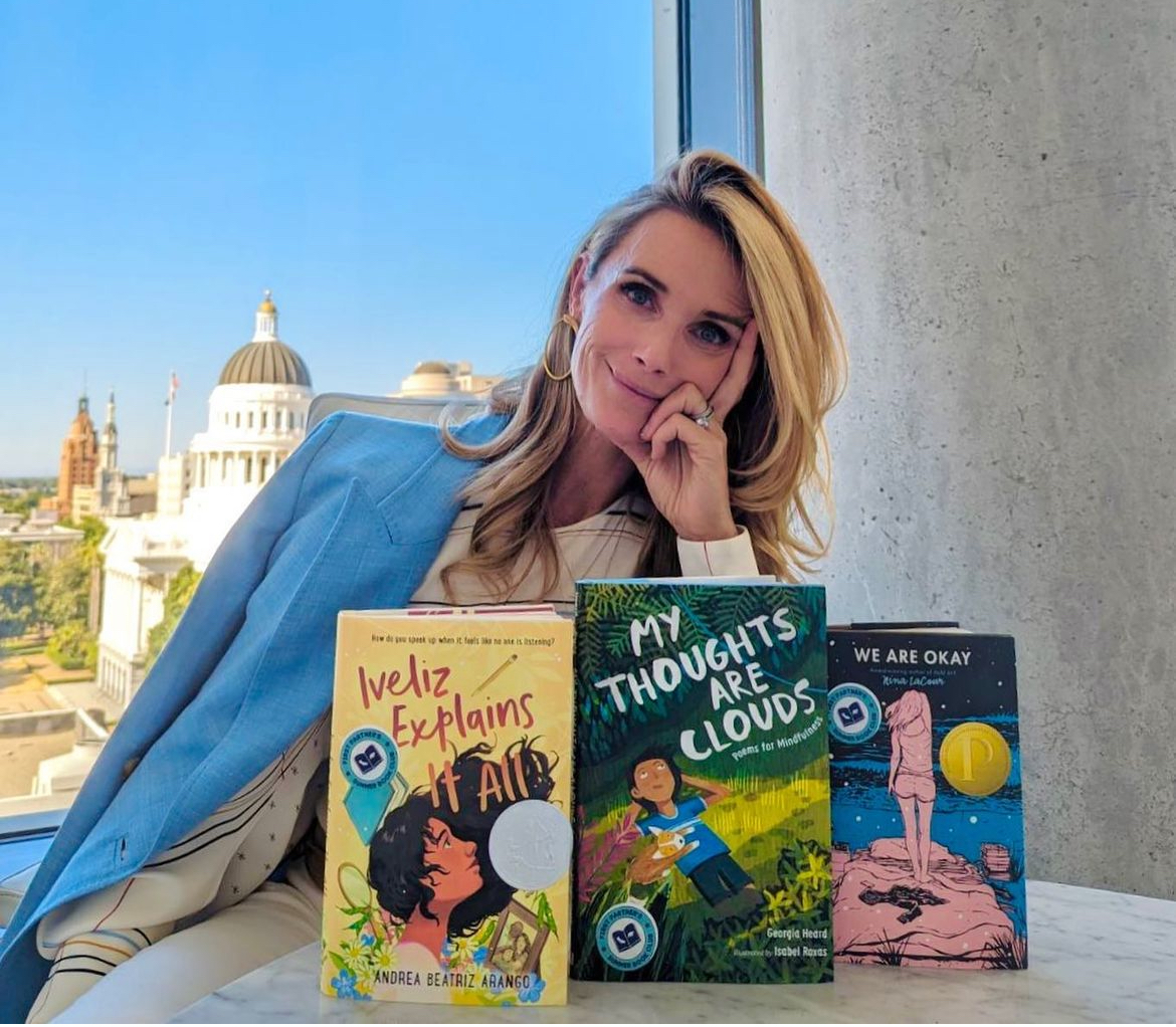 First Partner Jennifer Siebel Newsom smiling as she rests her head on her hand smiling. She is sitting in front of 3 books from her book list.