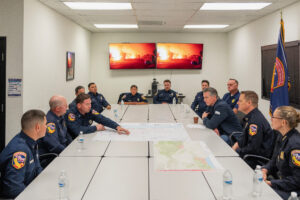 A photo of a large table with multiple firefighters in uniforms showing maps of firelines to Governor Newsom.