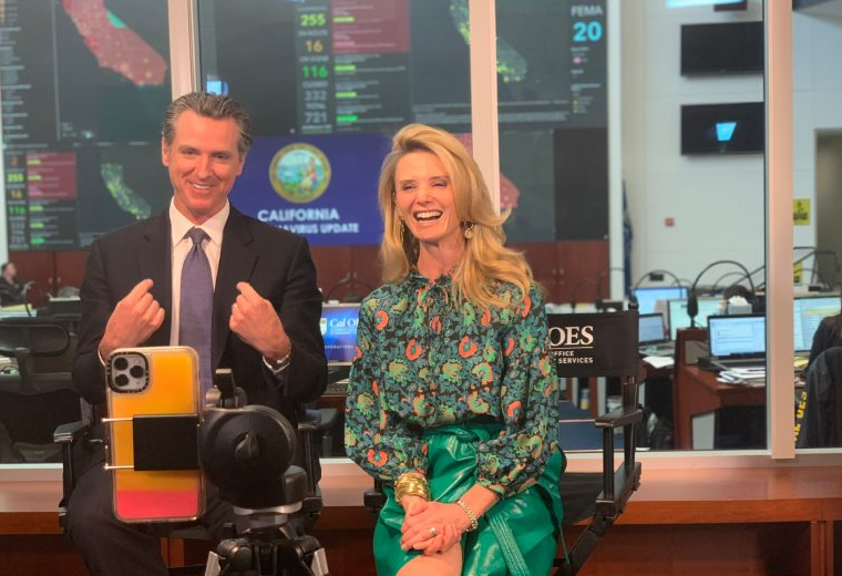 Governor Gavin Newsom, wearing a black suit with a periwinkle tie, sitting with First Partner Jennifer Siebel Newsom, who is wearing a green floral pattern blouse and green leather skirt. The two sit behind a cellphone propped by a tripod, smiling as they prepare to take a photo.