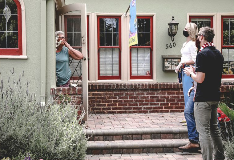 A blonde, light-skinned woman and a grey-haired, light-skinned man stand on the steps of a home that is painted light green with red window frames, both wearing masks. They are facing the front door, and are greeted by a grey-haired, light-skinned woman with glasses. The woman is gesturing an air kiss from the front door.